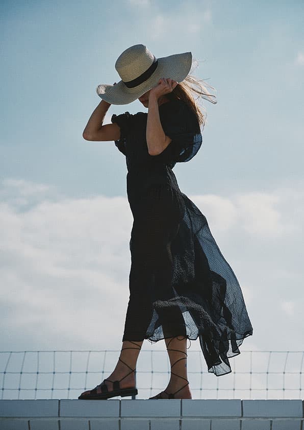 Une femme vêtue dune robe noire fluide, des sandales noires et dun chapeau à larges bords se tient debout sur un rebord contre un ciel nuageux.