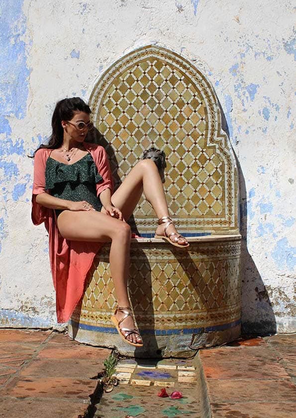 A woman wearing sunglasses, a forest green bodysuit, a light orange kimono and gold pink sandals sits on an ornate tiled fountain against a white and blue patina wall, with one leg raised, in the sun outside. Folegandros sandals.