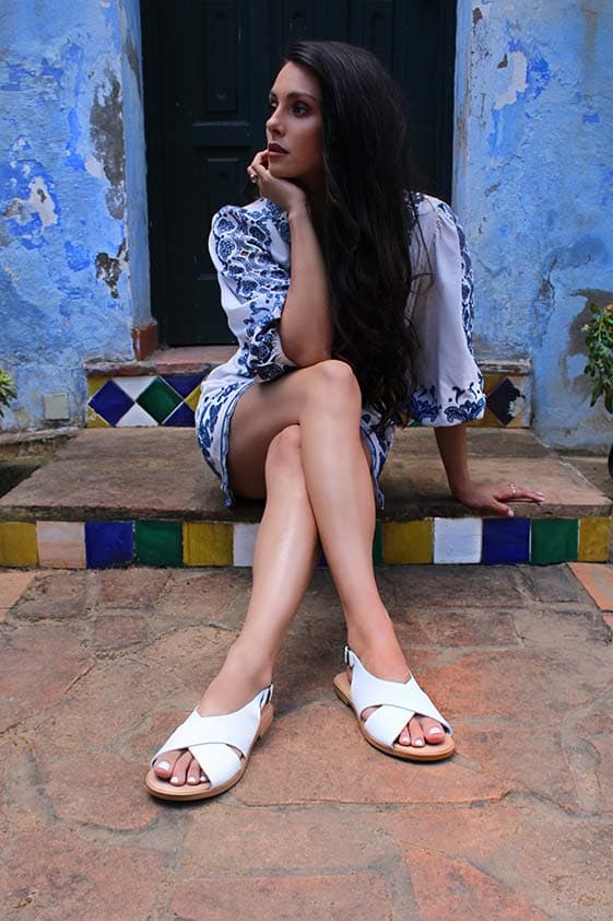 Woman in white dress with blue pattern and white leather sandals, sitting on steps, chin resting on one hand. Background: blue wall, green door, edge of tiled steps. Kriti sandals.