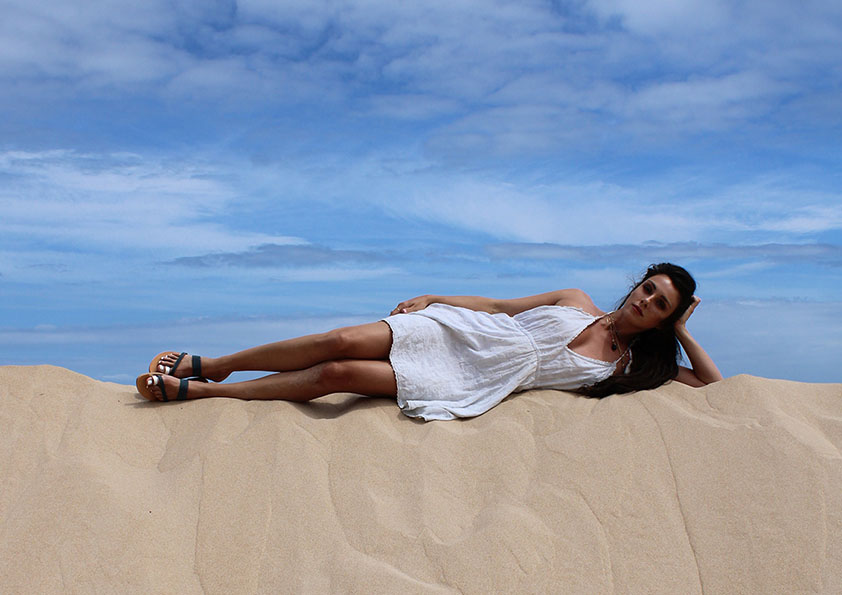 Woman in white dress lying on a sand dune under a blue sky with clouds, her hand supporting her head, her legs stretched out, wearing silver and grey leather sandals. Epidavros sandals.
