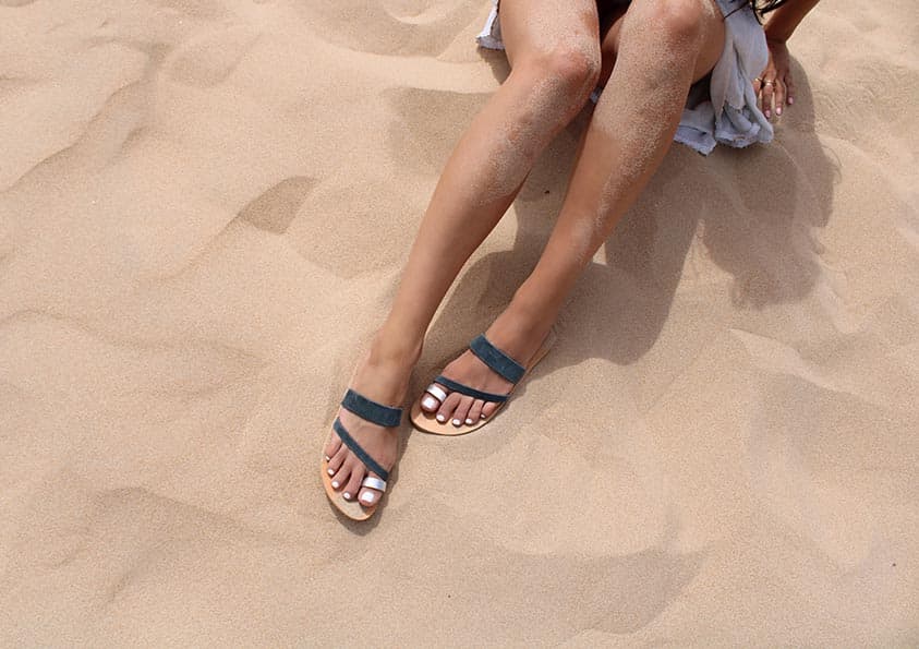 Personne assise sur une plage de sable, portant des nu-pieds en cuir gris et argenté. Jambes tendues, le sable sur la peau. Sandales Epidavros.
